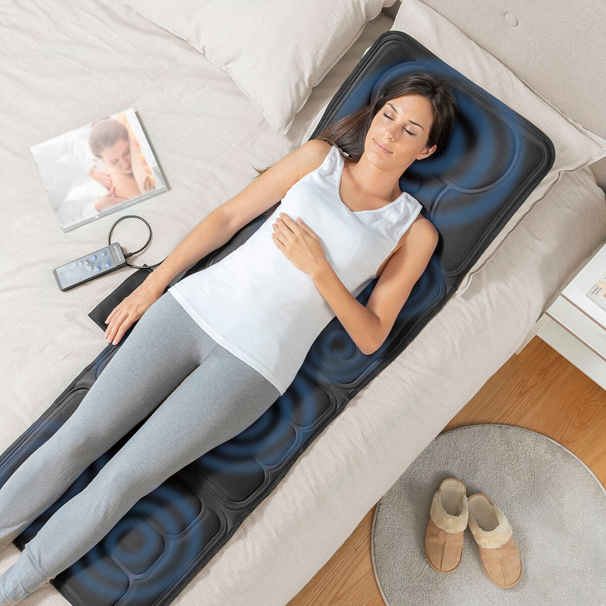 Mujer relajándose en la esterilla de masaje corporal InnovaGoods sobre una cama, con control remoto a su lado.