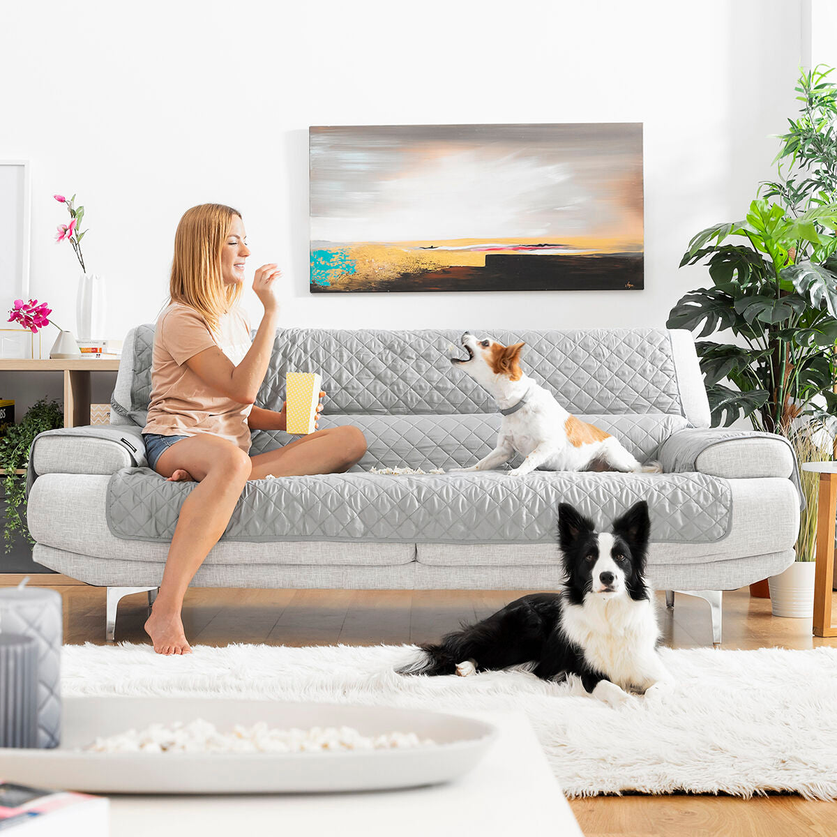 Reversible sofa protector cover on a modern couch with a woman and two playful dogs enjoying snacks at home.